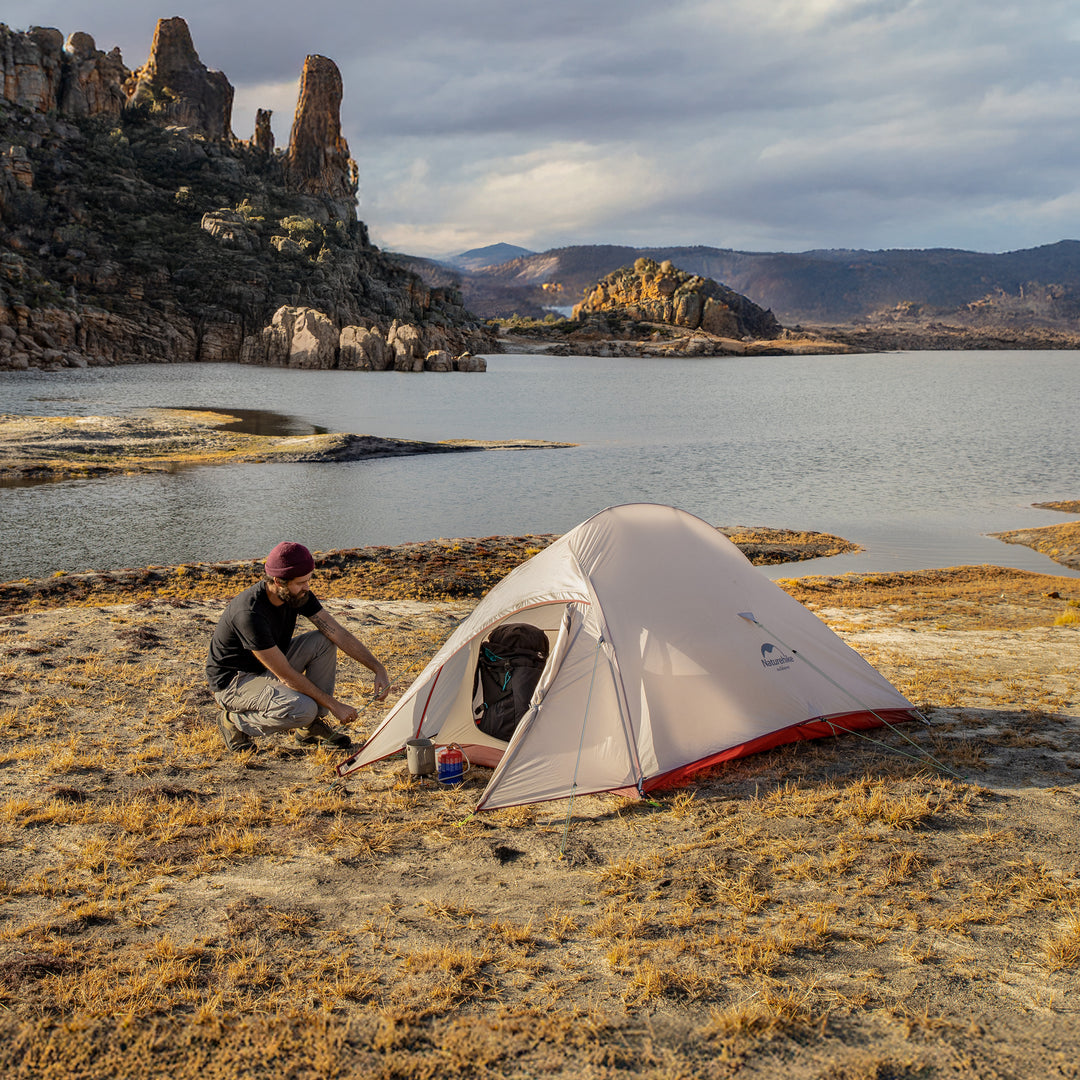 Naturehike Cloud Up Pro 3-Person ultralight backpacking tent set up by a mountain lake, with a person assembling the tent and a backpack inside, waterproof and wind-resistant shelter for hiking and camping.