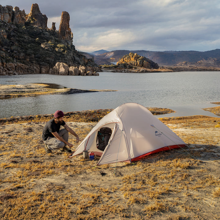 Naturehike Cloud Up Pro 3-Person ultralight backpacking tent set up by a mountain lake, with a person assembling the tent and a backpack inside, waterproof and wind-resistant shelter for hiking and camping.