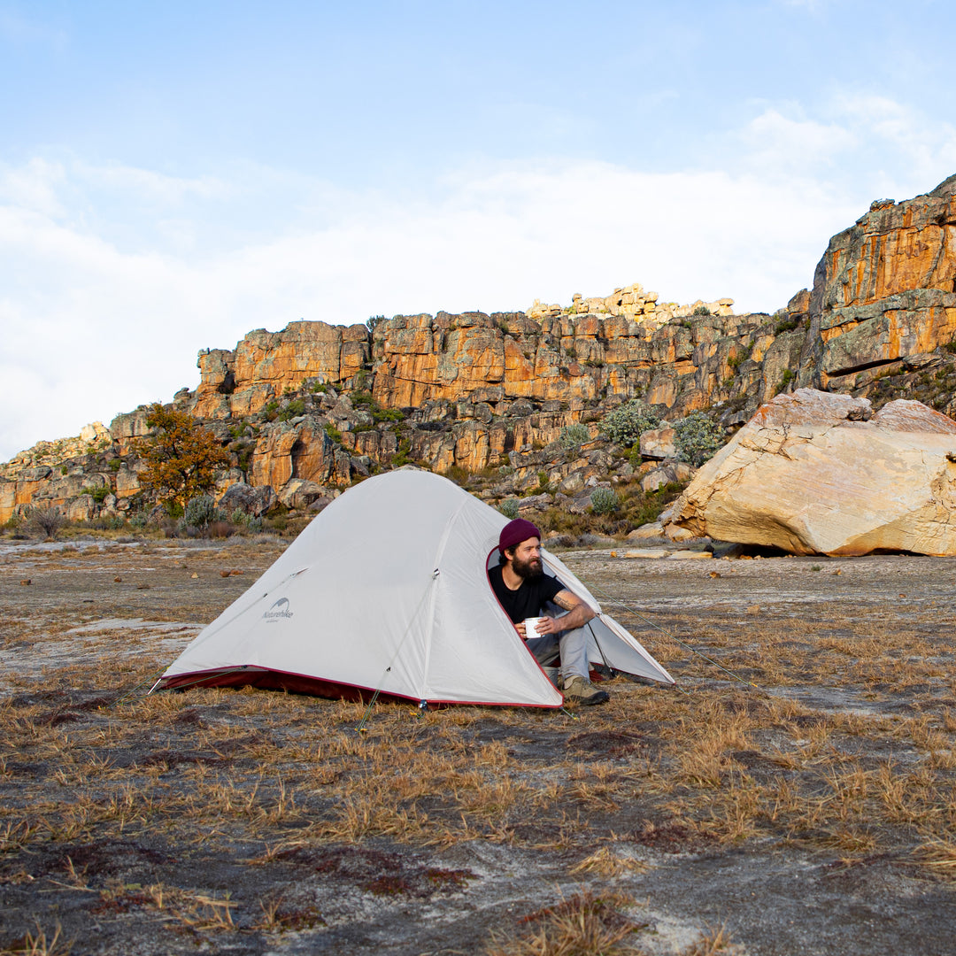 A person sitting in the vestibule of the Naturehike Cloud Up Pro 3-Person ultralight backpacking tent outdoors, enjoying coffee, showing spacious and comfortable camping setup.