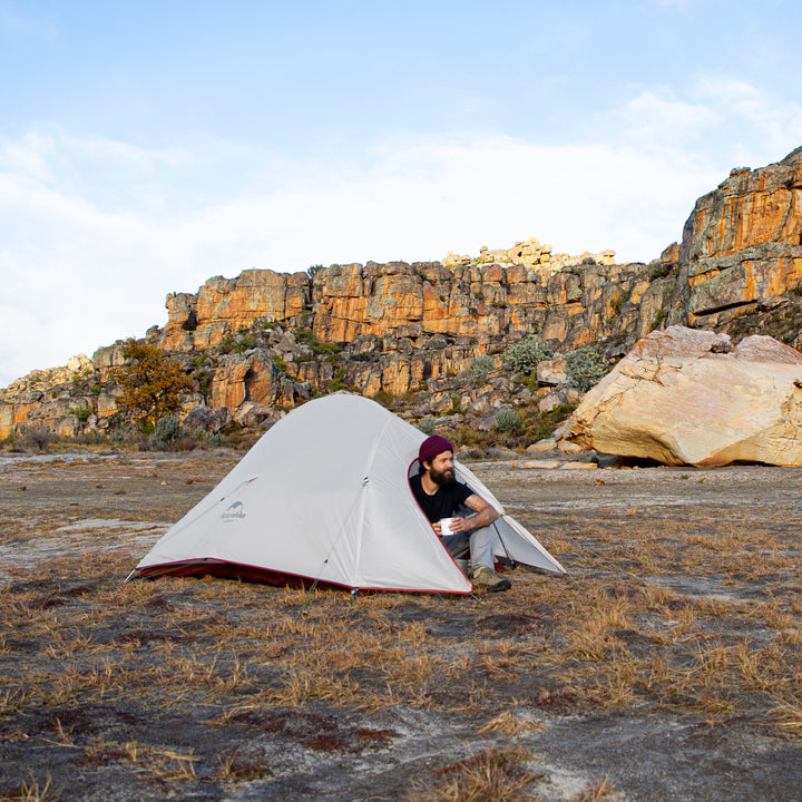 A person sitting in the vestibule of the Naturehike Cloud Up Pro 3-Person ultralight backpacking tent outdoors, enjoying coffee, showing spacious and comfortable camping setup.