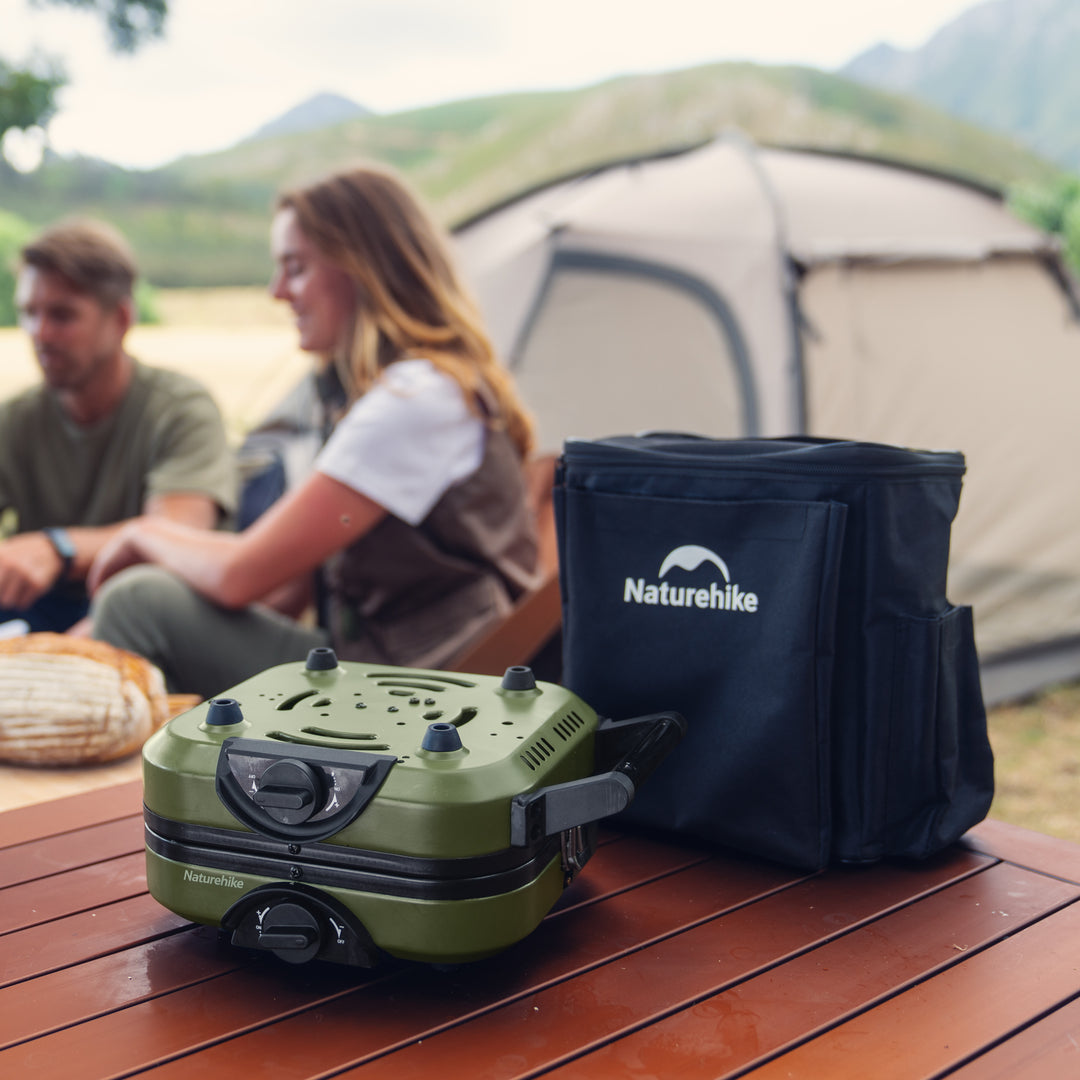 The Naturehike Phantom 2-Burner Folding Camping Stove packed neatly in its carry bag, resting on a camping table, emphasizing its small footprint and easy storage among other essential camp gear.